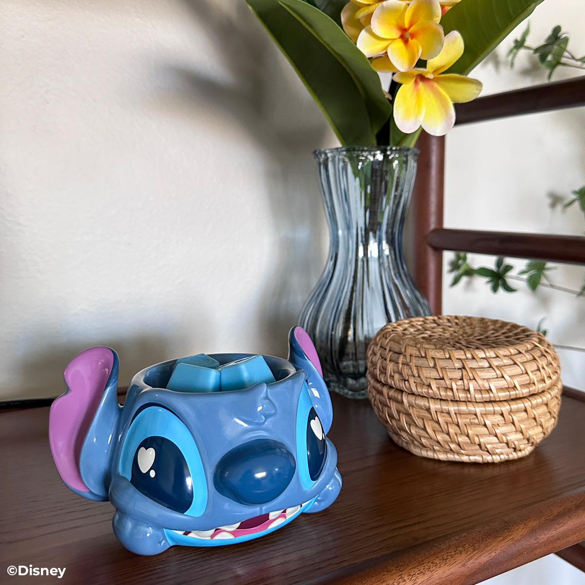 Blue character mug with pink accents on a wooden surface next to a vase with flowers and a woven basket.