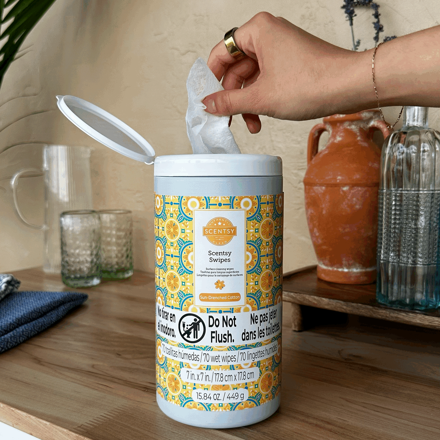 Hand pulling a wipe from a container of Scentsy Swipes on a wooden surface with decorative items in the background.