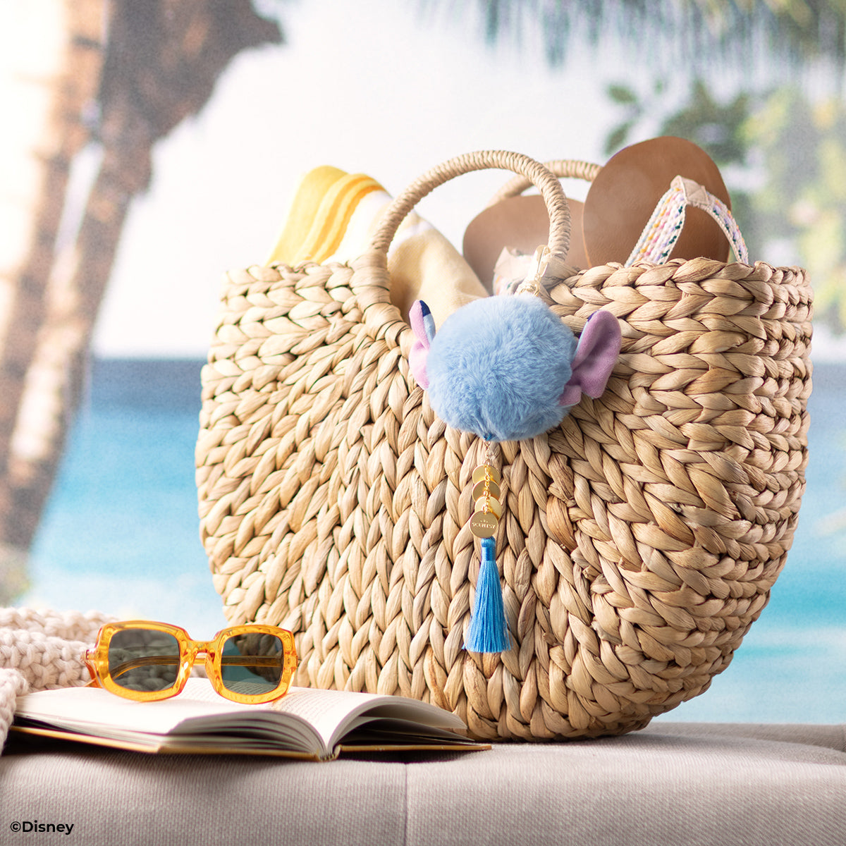 Woven bag with pom-pom and tassel, sunglasses, and book on a beach setting