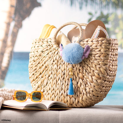Woven bag with pom-pom and tassel, sunglasses, and book on a beach setting