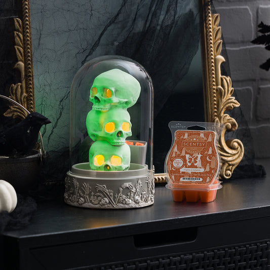 Green skeleton figurine under a glass dome on a dark surface with a Scentsy wax melt container.