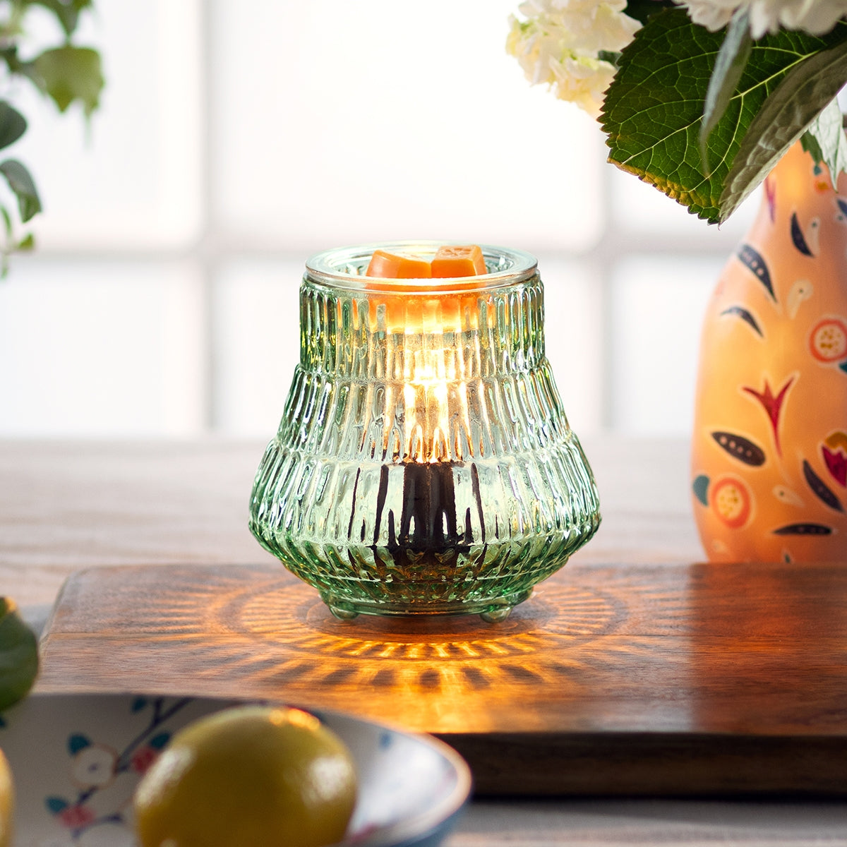 Green glass candle holder with a lit candle on a wooden surface, next to a colorful vase and lemons.