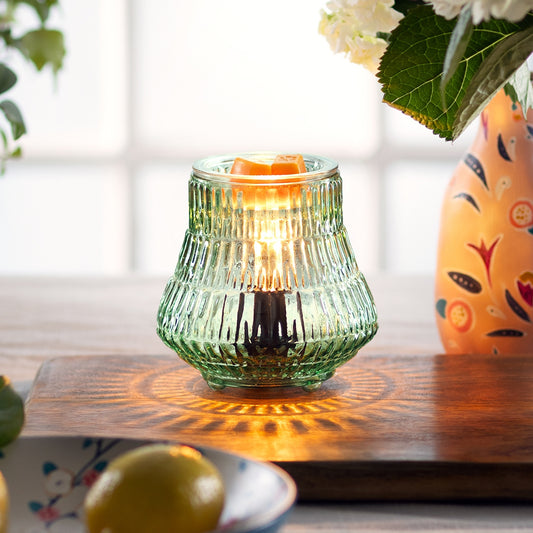 Green glass candle holder with a lit candle on a wooden surface, next to a colorful vase and lemons.