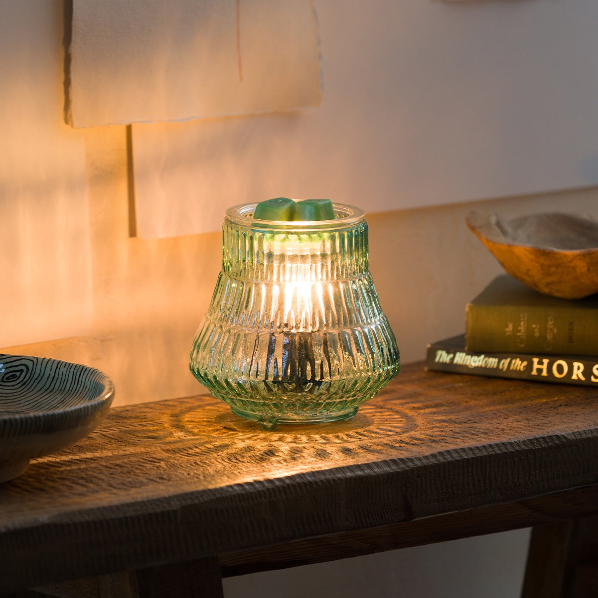 Green glass candle holder with a lit candle on a wooden surface, with books and a bowl in the background.